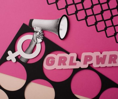 A creative and bold flat lay artwork featuring feminist symbols on a pink background. A hand holding a megaphone emerges from a female gender symbol cutout, and the phrase “GRL PWR” (Girl Power) is displayed in large pink letters. The design includes a mix of black and pink geometric patterns, emphasizing empowerment and modern feminism.