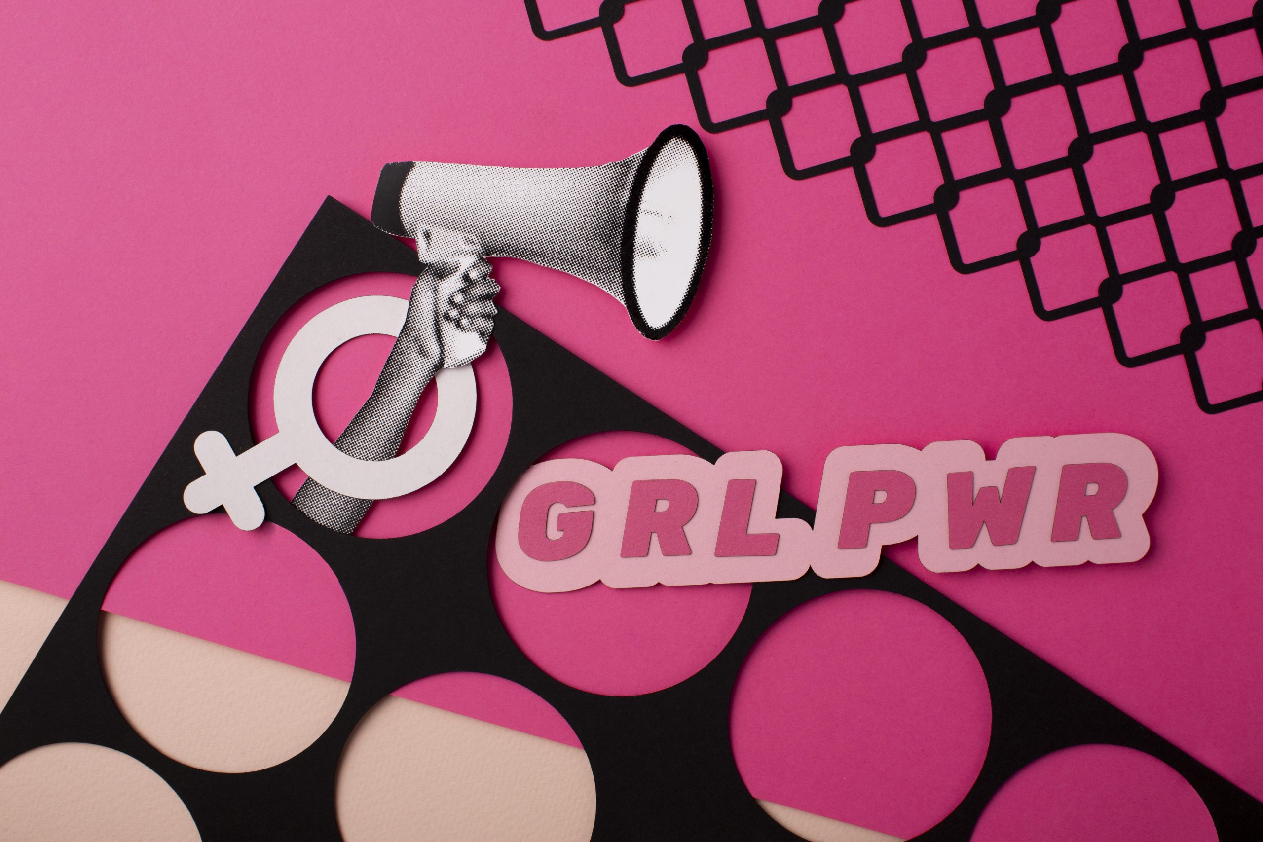 A creative and bold flat lay artwork featuring feminist symbols on a pink background. A hand holding a megaphone emerges from a female gender symbol cutout, and the phrase “GRL PWR” (Girl Power) is displayed in large pink letters. The design includes a mix of black and pink geometric patterns, emphasizing empowerment and modern feminism.